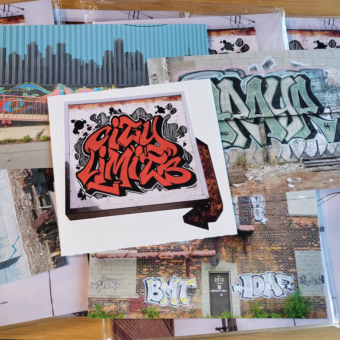 A grouping of small magazines wrapped in plastic behind photo copies of graffiti pieces from various artists with a sticker on top of the pile reading ' City Limits' written in red graffiti style letters, pasted on a cropped out photo of an old backlit sign with a rusted arrow at the right side