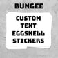 A stack of white eggshell-shaped stickers with bold black text reading 'BUNGEE CUSTOM TEXT EGGSHELL STICKERS' on a textured gray background.