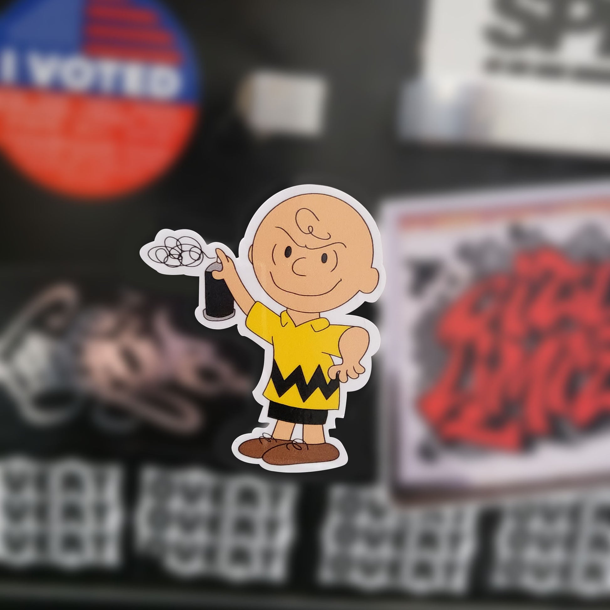 A die-cut sticker of the cartoon character Charlie Brown from Peanuts, shown against a blurred background of other stickers and magnets.