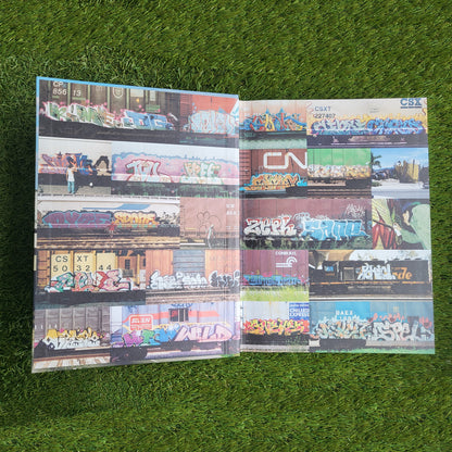 Open hardcover book, the inner cover shows a collage of photos of trains with graffiti writing covering both sides of the pages. Background is a green grass