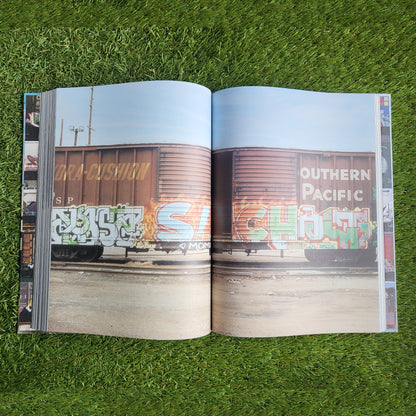 An open, hardcover book showing a southern pacific train with graffiti writing on it covering both pages. Behind the book is a grass background