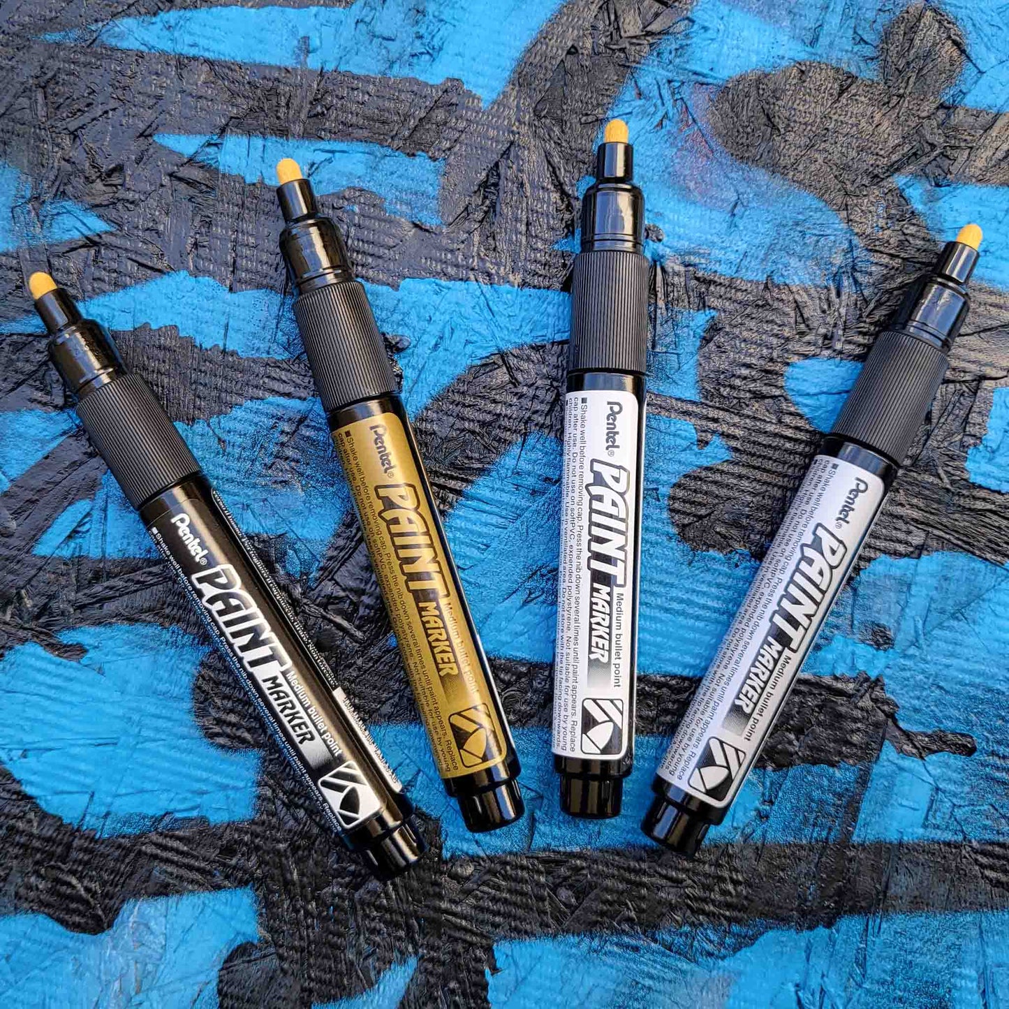 Four pentel paint markers in black, gold, silver and white on a textured blue and black background
