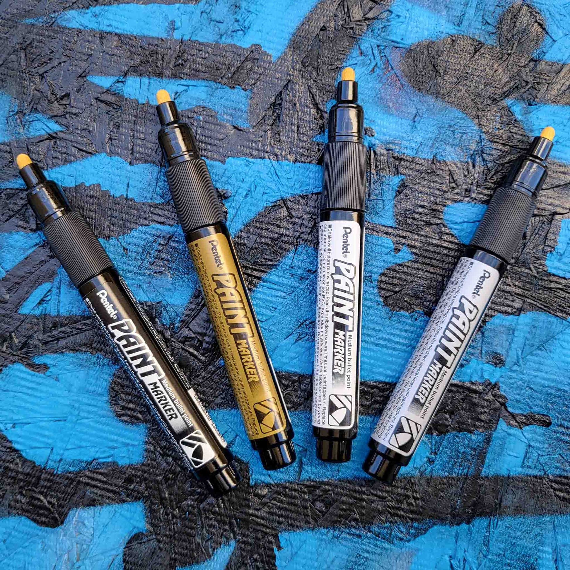Four pentel paint markers in black, gold, silver and white on a textured blue and black background
