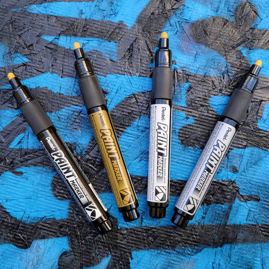 Four pentel paint markers in black, gold, silver and white on a textured blue and black background