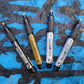 Four pentel paint markers in black, gold, silver and white on a textured blue and black background