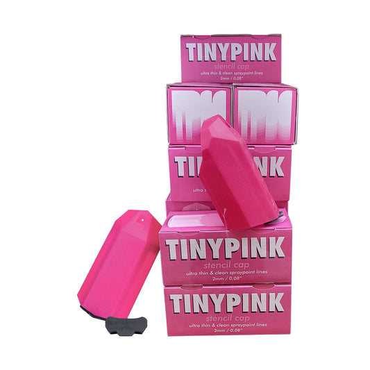 A stack of pink and white small boxes on the right side read "TINYPINK - stencil cap - ultra thing & clean spraypaint lines" in white. The boxes are offput each level, there are two boxes stacked in front of the pile with a hot pink, plastic adapter that has a small hole close to the top. There is another adapter to the left that is leaning on the boxes to the right.