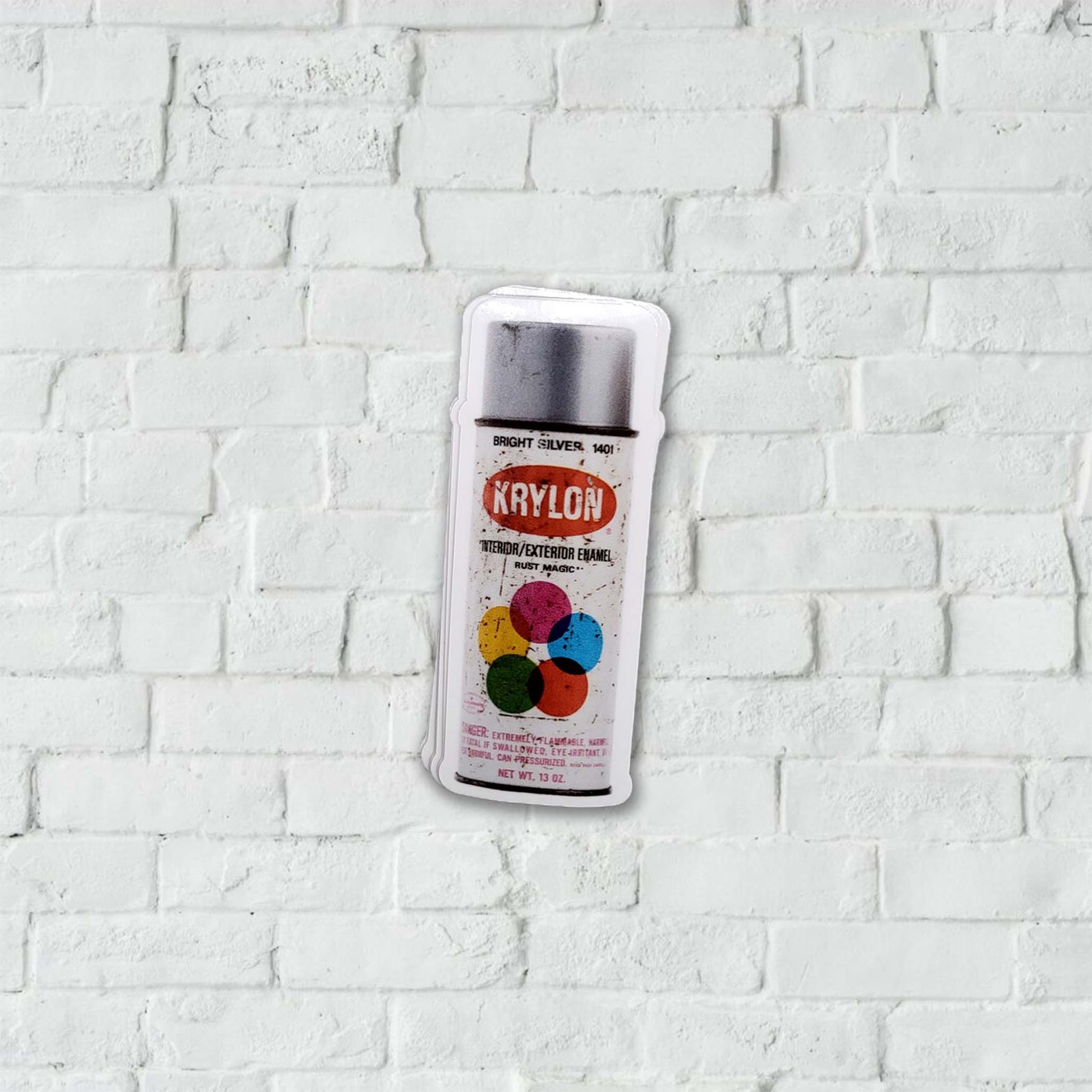 Silver Krylon spray paint can stickers on a white brick wall background.