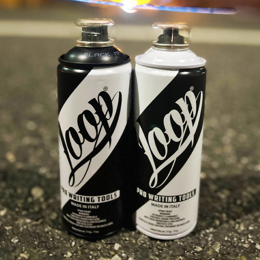 Two spray paint cans, on the left a black can with a white, thick stripe running diagonally across the face with the logo "Loop" written in cursive. The right, a white can with a black, thick stripe wrapping across diagonally with the word "Loop" written on the face in script. The background is asphalt with a blurry light at the top.
