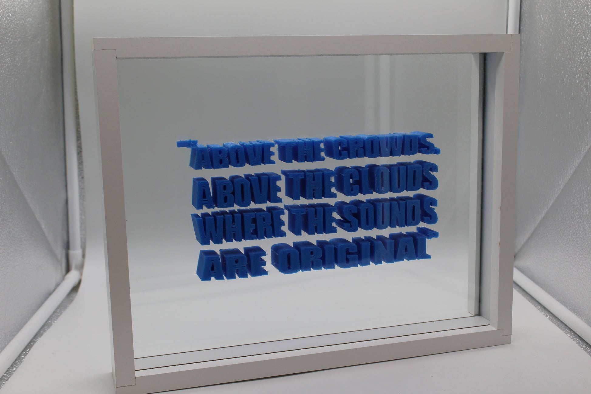 White box framing a glass sheet with blue plastic letters in between the slates of glass, background is in a small white and silver photobox, it reads , '"Above the crowds, above the clouds, where the sounds are original" in capital letters